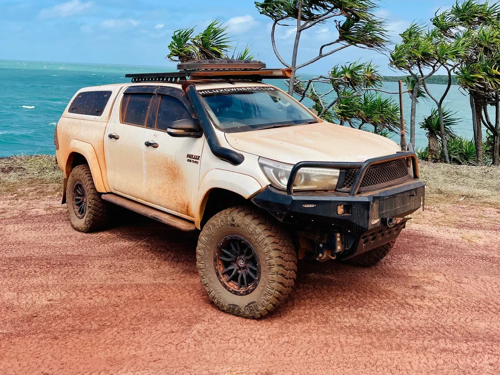 A dusty Hilux on red dirt above the sea, pandanus palms behind.