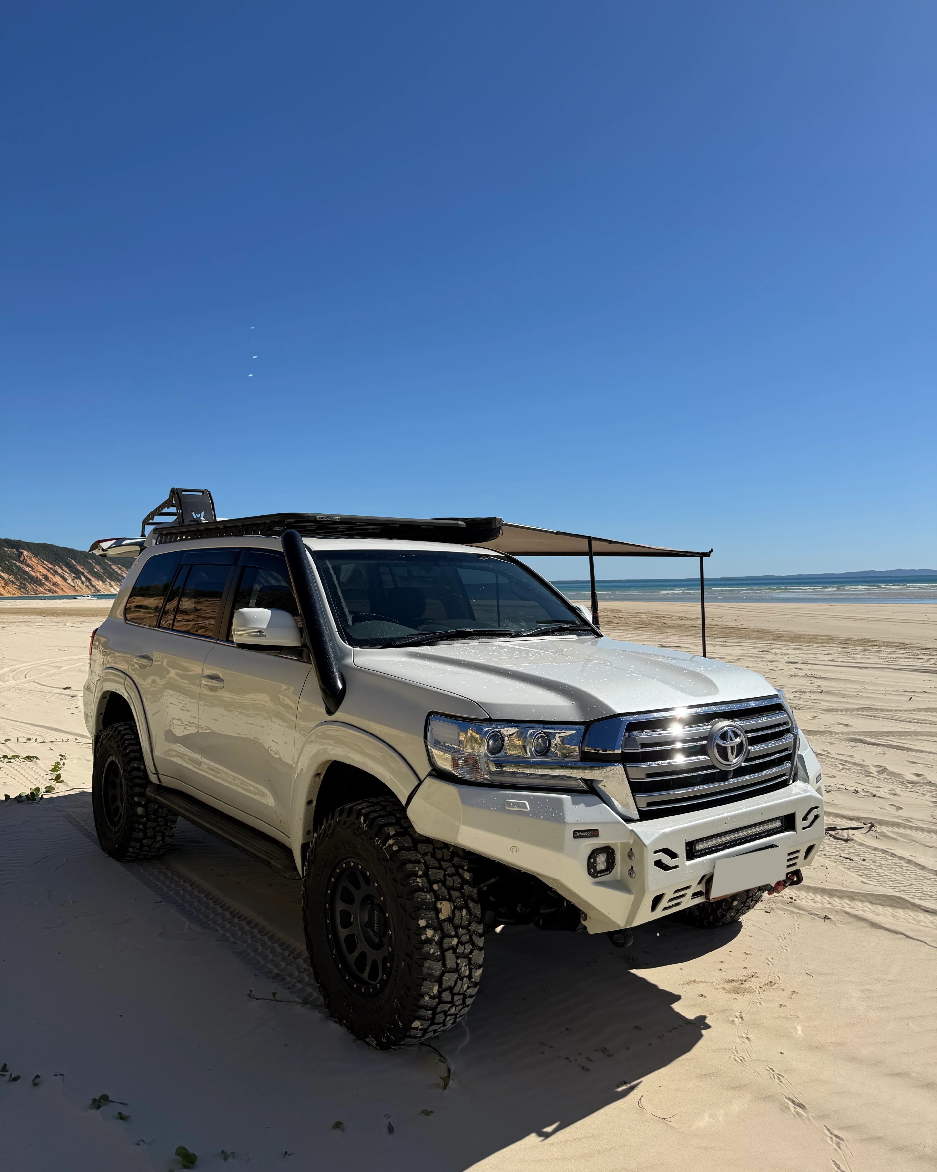 A white 200 Series on the beach with the awning out and a roof rack loaded.