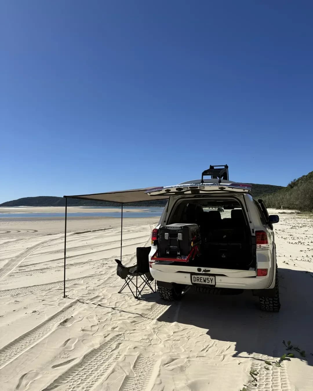 A 200 Series on the sand with the rear open, awning out and a camp chair beside it.