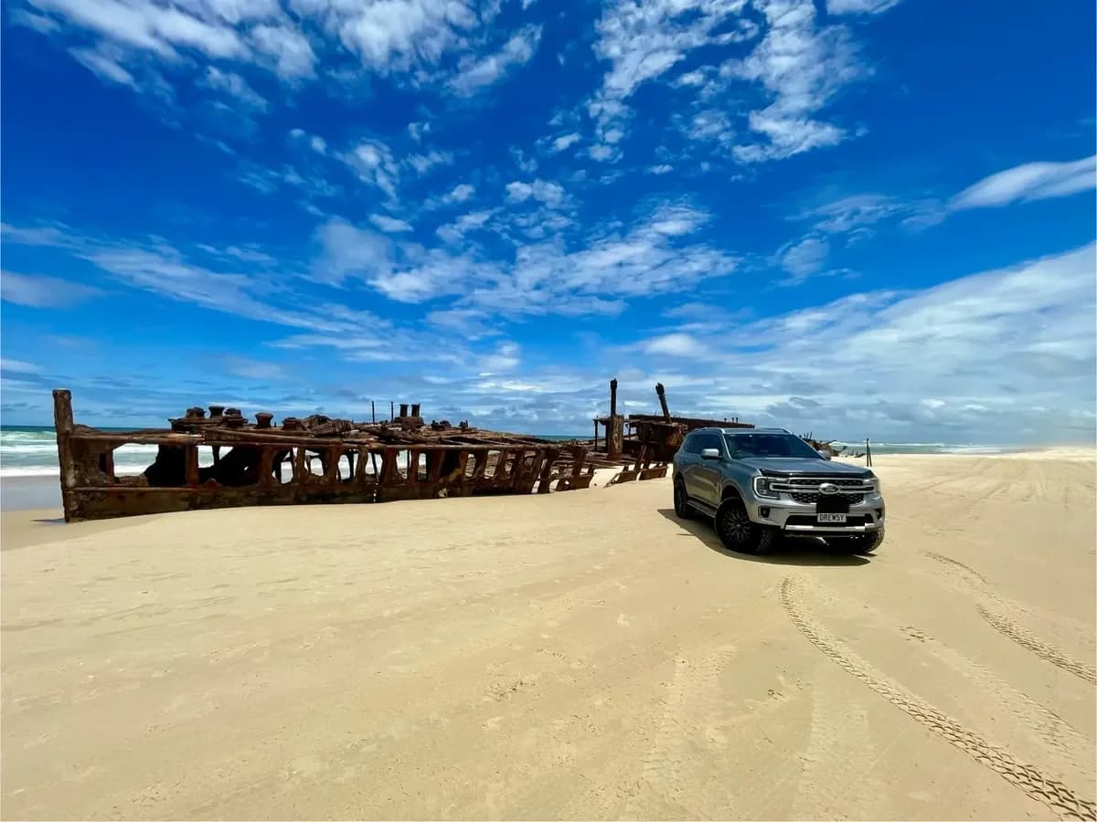 A 4x4 parked on a wide beach beside a rusted shipwreck.