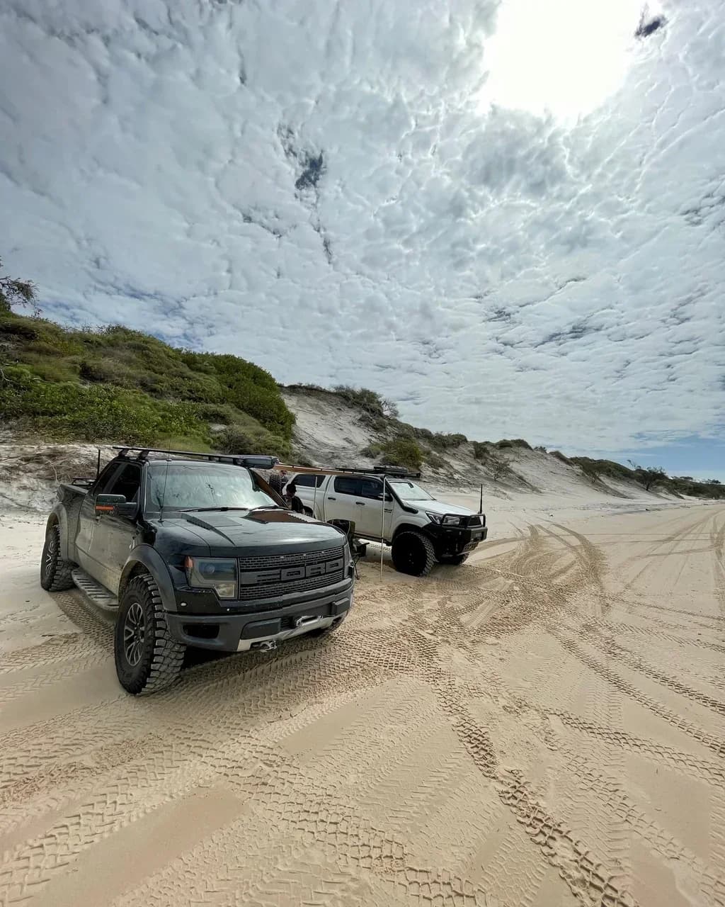 A Ford Raptor and a Hilux parked on a wide beach below vegetated dunes under a big sky.