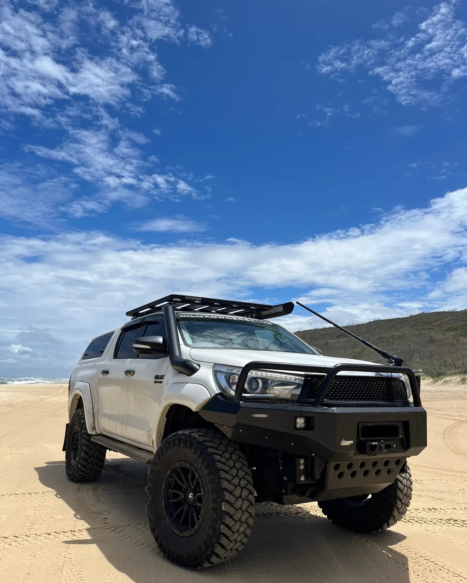 A white Hilux with a bull bar, snorkel and roof rack on a wide beach under a blue sky.