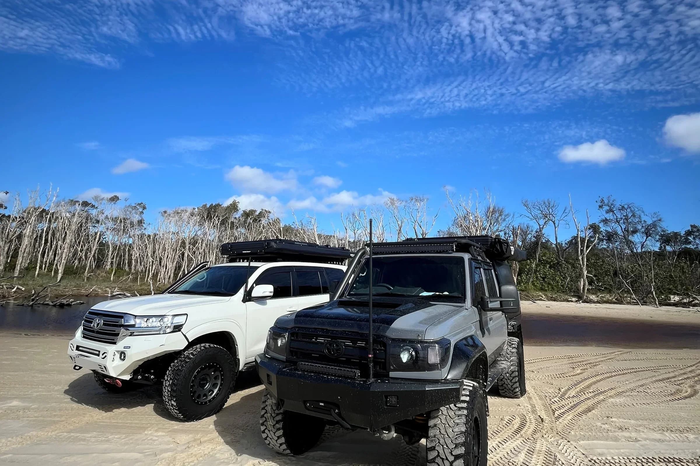 Drewsy's 79 and a white 200 Series side by side on the sand under a big mackerel sky.
